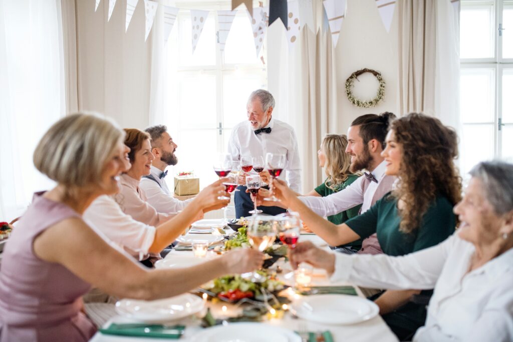 A family at the dining room table during the holiday season. An older man at the end of the table is standing and offering a toast.
