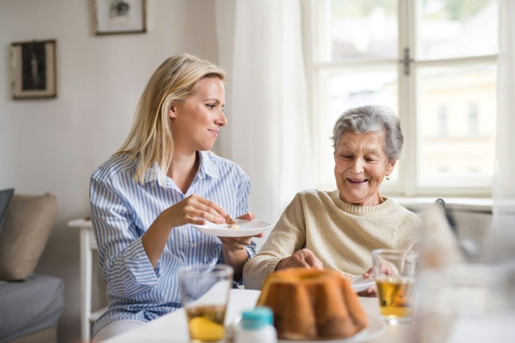 Two women in a retirement home in front of a window, drinking iced tea and eating pound cake. One is the adult daughter of the other woman.