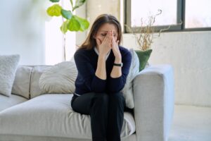 A woman sits on a couch with her face in her hands, frustrated, sad, burned out.